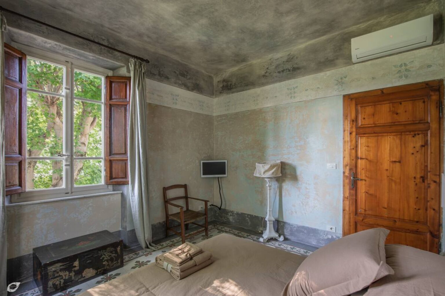 Spacious bedroom with rustic decor, featuring a wooden door, a chair, and a small lamp beside the bed, with large windows allowing natural light and a view of greenery outside. | Only on SopranoVillas
