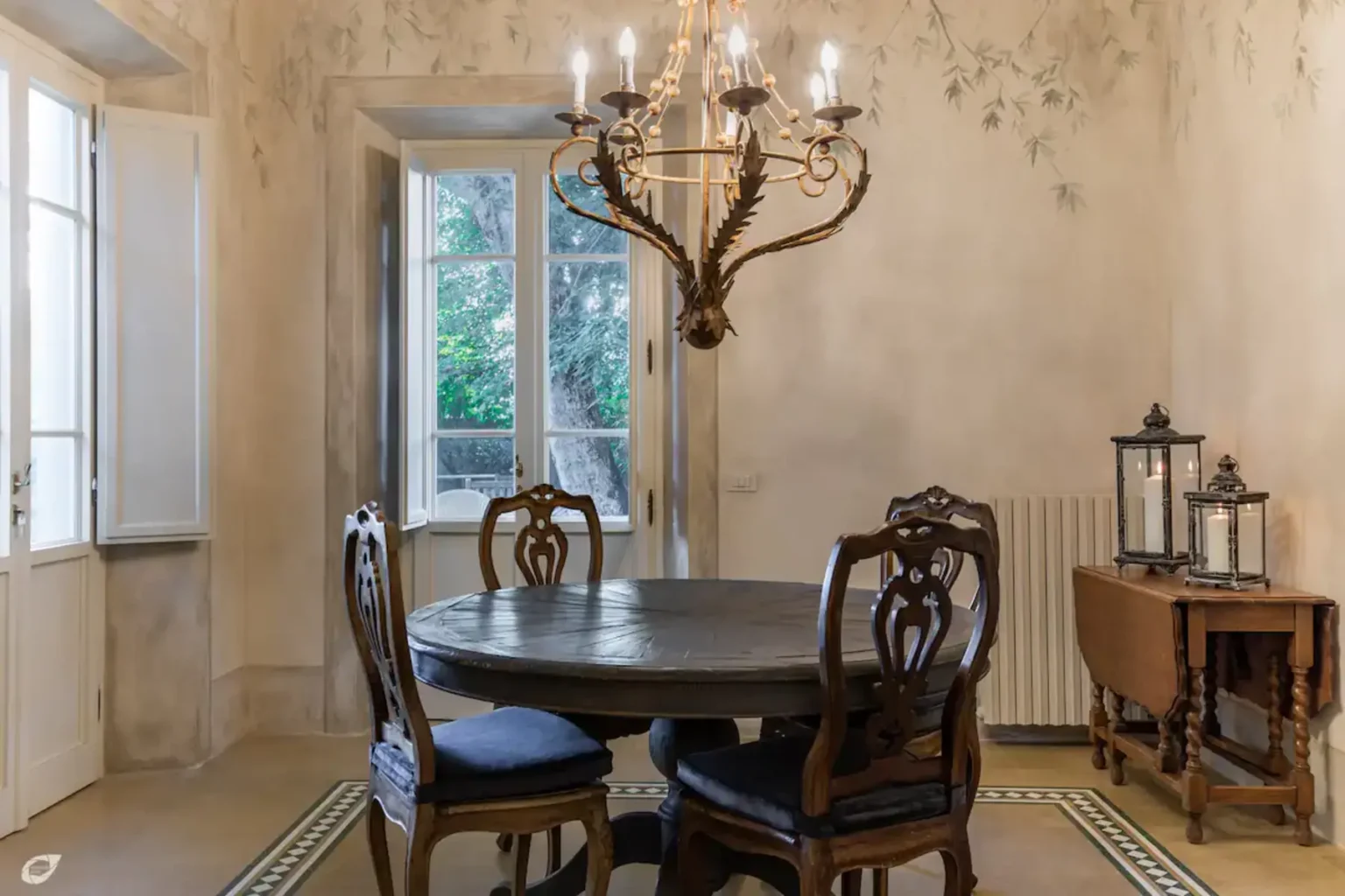 Elegant dining room featuring a round wooden table, antique chairs with blue upholstery, a vintage side table, and a chandelier, illuminated by natural light from large windows. | Only on SopranoVillas