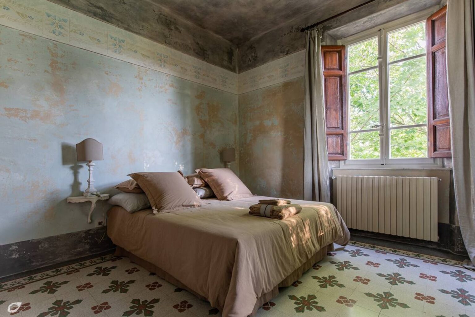 Cozy bedroom featuring a double bed with beige linens and decorative pillows, a bedside lamp on a wall-mounted shelf, and a large window with wooden shutters allowing natural light. The room has vintage-style decor, including a patterned floor and soft curtains, creating a serene atmosphere. | Only on SopranoVillas