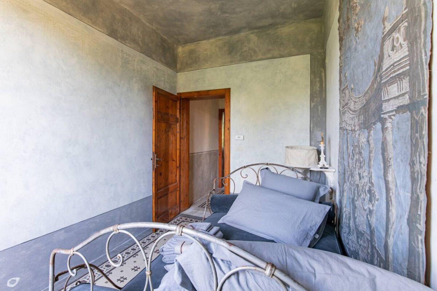 Cozy bedroom featuring a vintage metal bed with gray bedding, a wooden door, and decorative wall art, set in a minimalist design with soft lighting. | Only on SopranoVillas