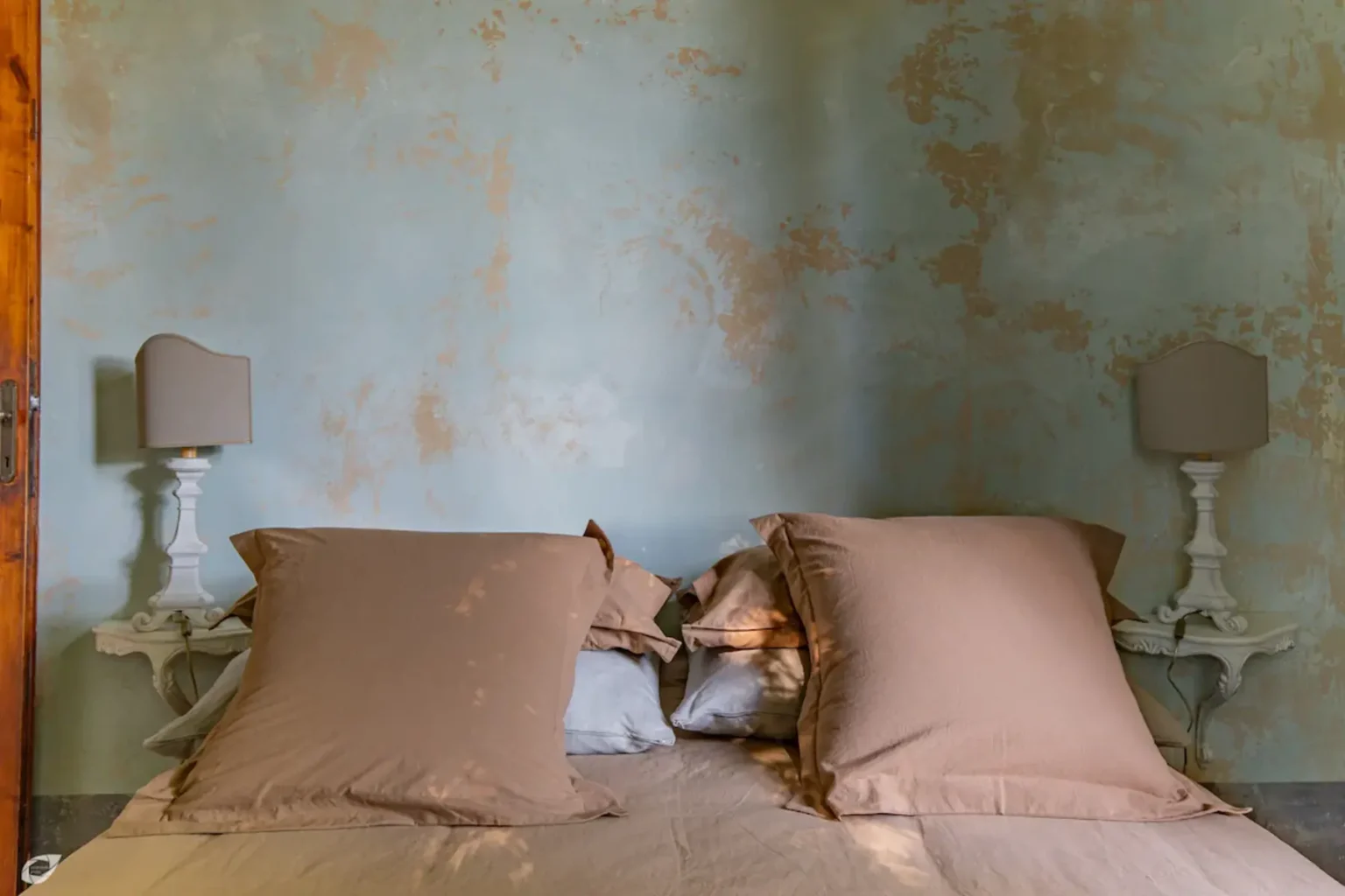 Cozy bedroom interior featuring a bed with large brown pillows, soft bedding, and vintage lamps on either side against a textured blue wall. | Only on SopranoVillas