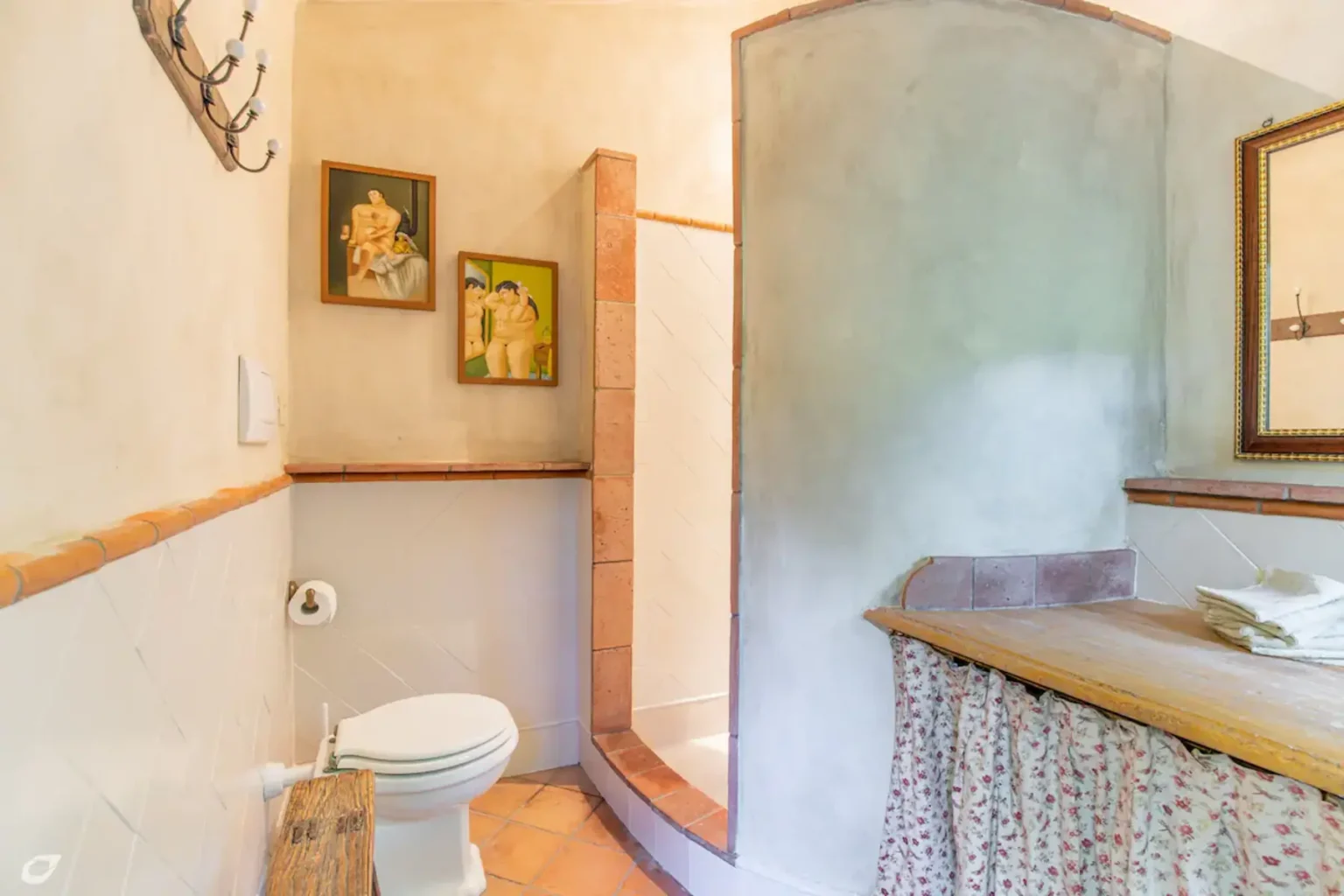 Cozy bathroom featuring rustic decor, including framed artwork, a wooden countertop with floral curtains, and a shower area. The space is illuminated by natural light, highlighting the textured walls and terracotta tiles. | Only on SopranoVillas
