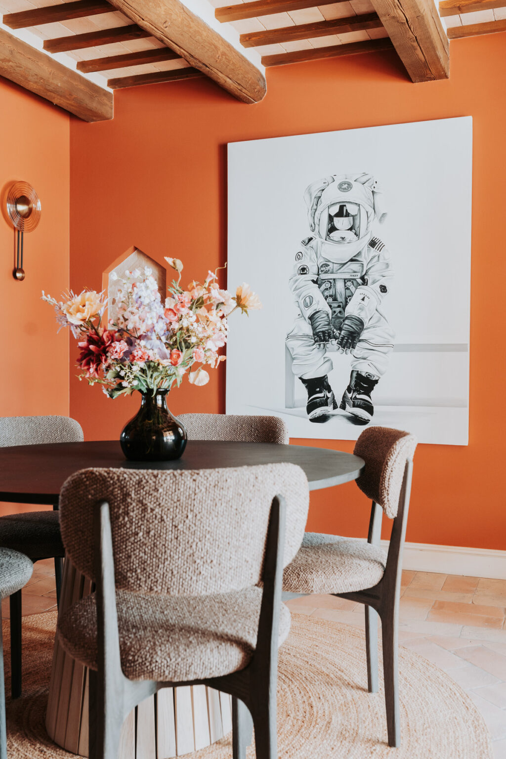 Stylish dining area featuring a round table with textured chairs, adorned with a black vase of colorful flowers, set against a vibrant orange wall and a large astronaut-themed artwork. | Only on SopranoVillas