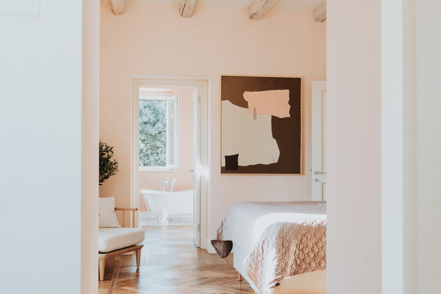 Modern bedroom interior featuring soft peach walls, a cozy bed with a quilt, and a contemporary art piece. A glimpse of a stylish sitting area and a bright bathroom is visible through the doorway. | Only on SopranoVillas