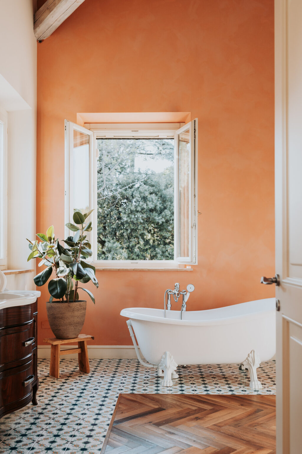 Bright bathroom featuring a freestanding white bathtub, an indoor plant, and a large window with a view of greenery, set against warm orange walls. Ideal for showcasing modern interior design and natural light in home decor. | Only on SopranoVillas