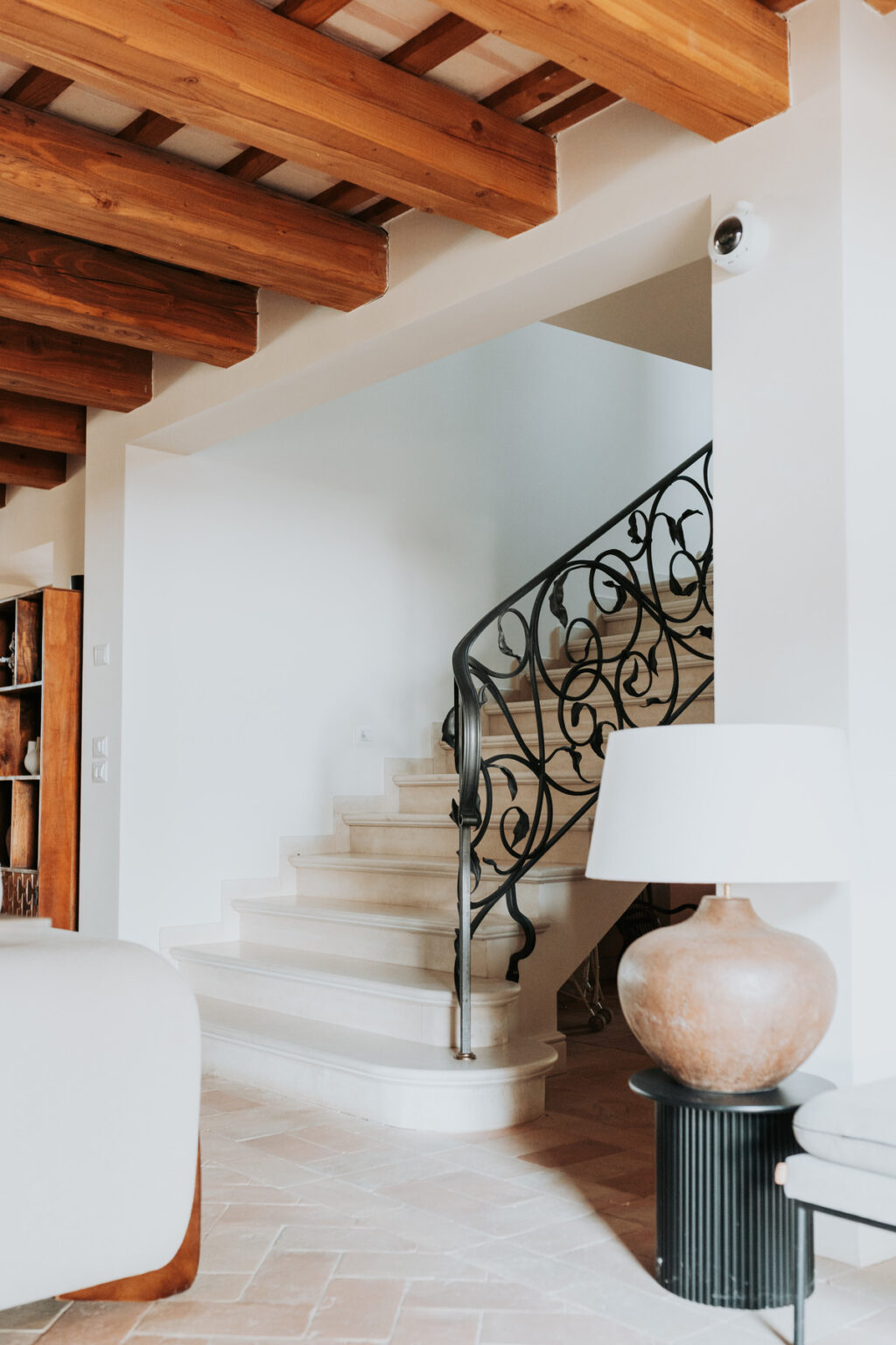 Elegant interior staircase with a decorative black wrought iron railing, leading to an upper level, complemented by a stylish lamp on a nearby table. Natural light enhances the warm, inviting atmosphere of the space. | Only on SopranoVillas