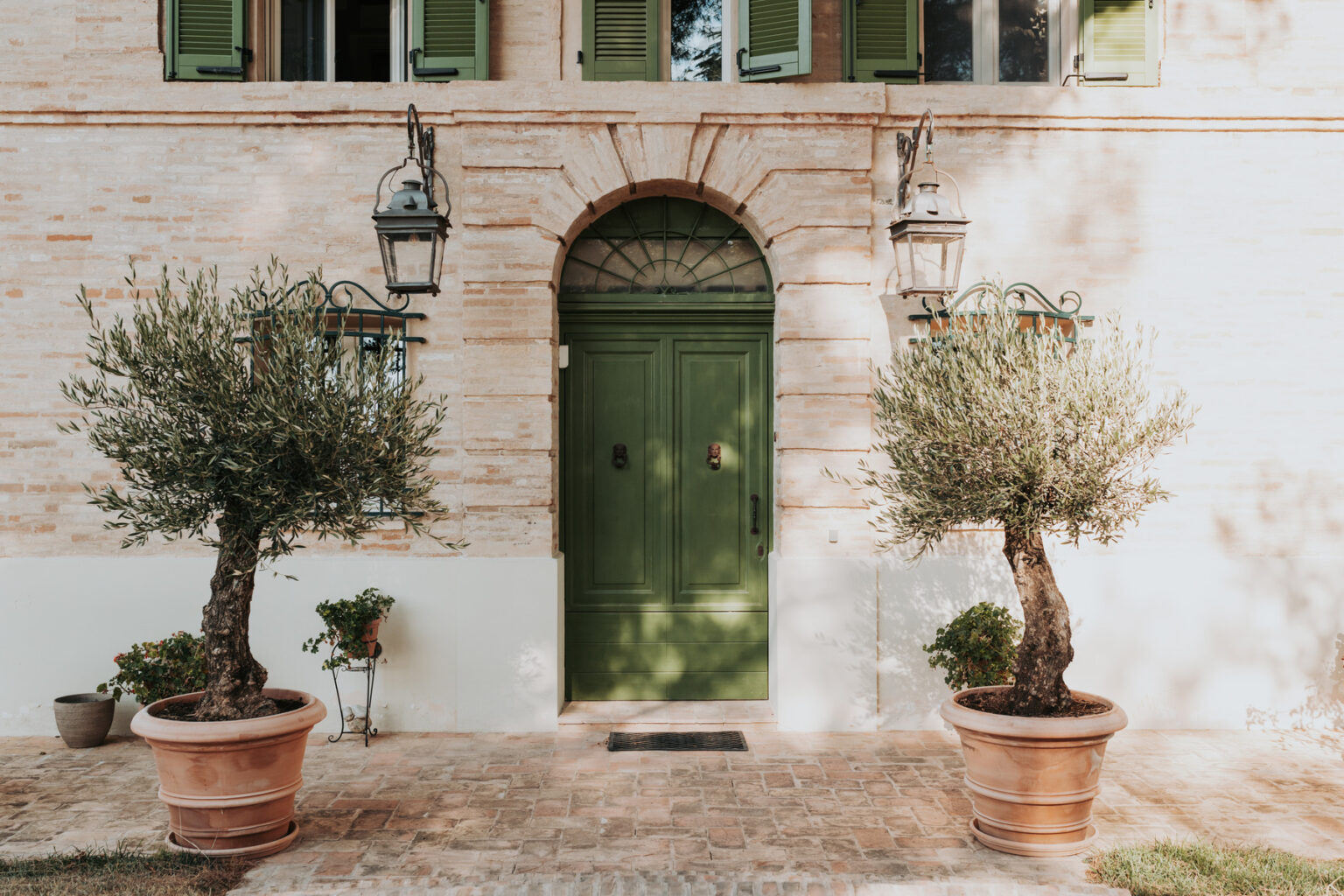 Charming entrance of a rustic building featuring a green door, flanked by two olive trees in terracotta pots and vintage lanterns hanging on either side. | Only on SopranoVillas