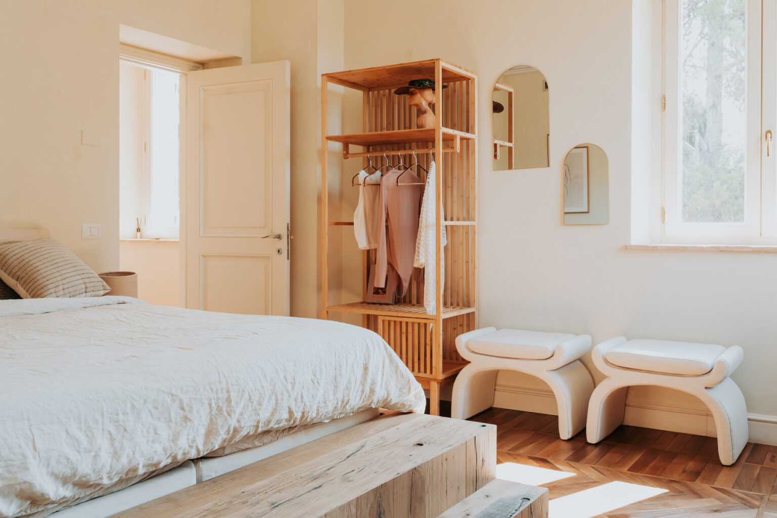 Cozy and modern bedroom featuring a wooden bed, minimalist decor, a stylish open wardrobe with hanging clothes, and two contemporary stools. Natural light streams in through large windows, enhancing the serene atmosphere. | Only on SopranoVillas
