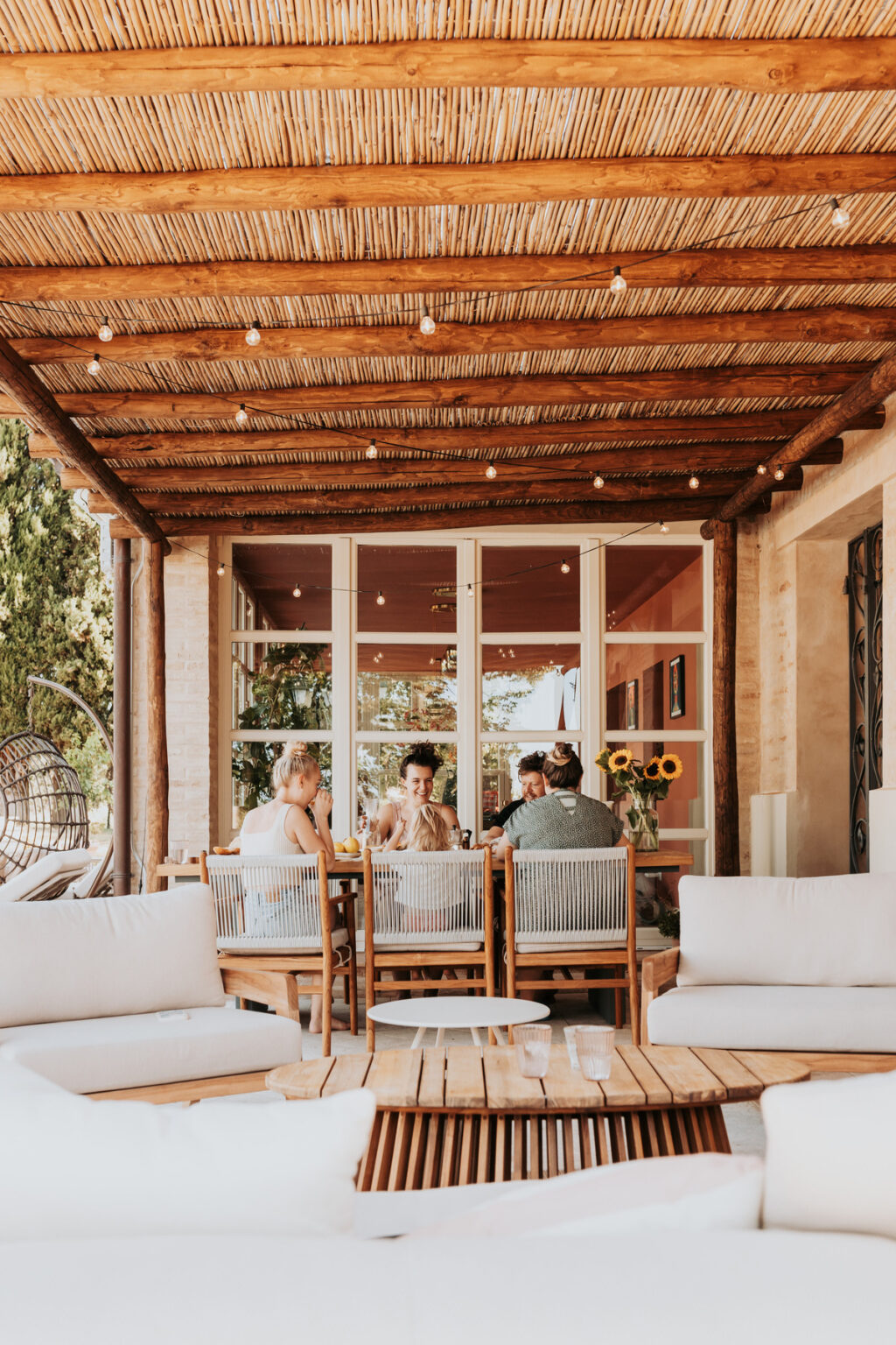 Outdoor dining area featuring a group of people enjoying a meal under a wooden pergola with string lights. The setting includes comfortable seating arrangements, a wooden table, and a backdrop of sunflowers and greenery, creating a warm and inviting atmosphere. | Only on SopranoVillas