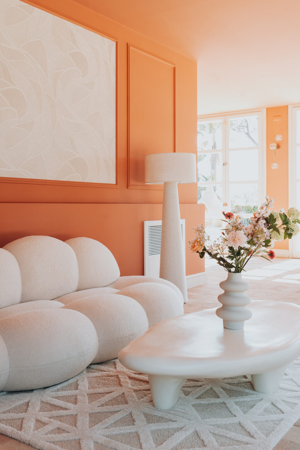 Modern living room featuring a plush white sofa, a sculptural white coffee table, and an elegant floor lamp against vibrant orange walls. A vase with fresh flowers adds a touch of color, complemented by a patterned area rug. Natural light floods the space through large windows. | Only on SopranoVillas