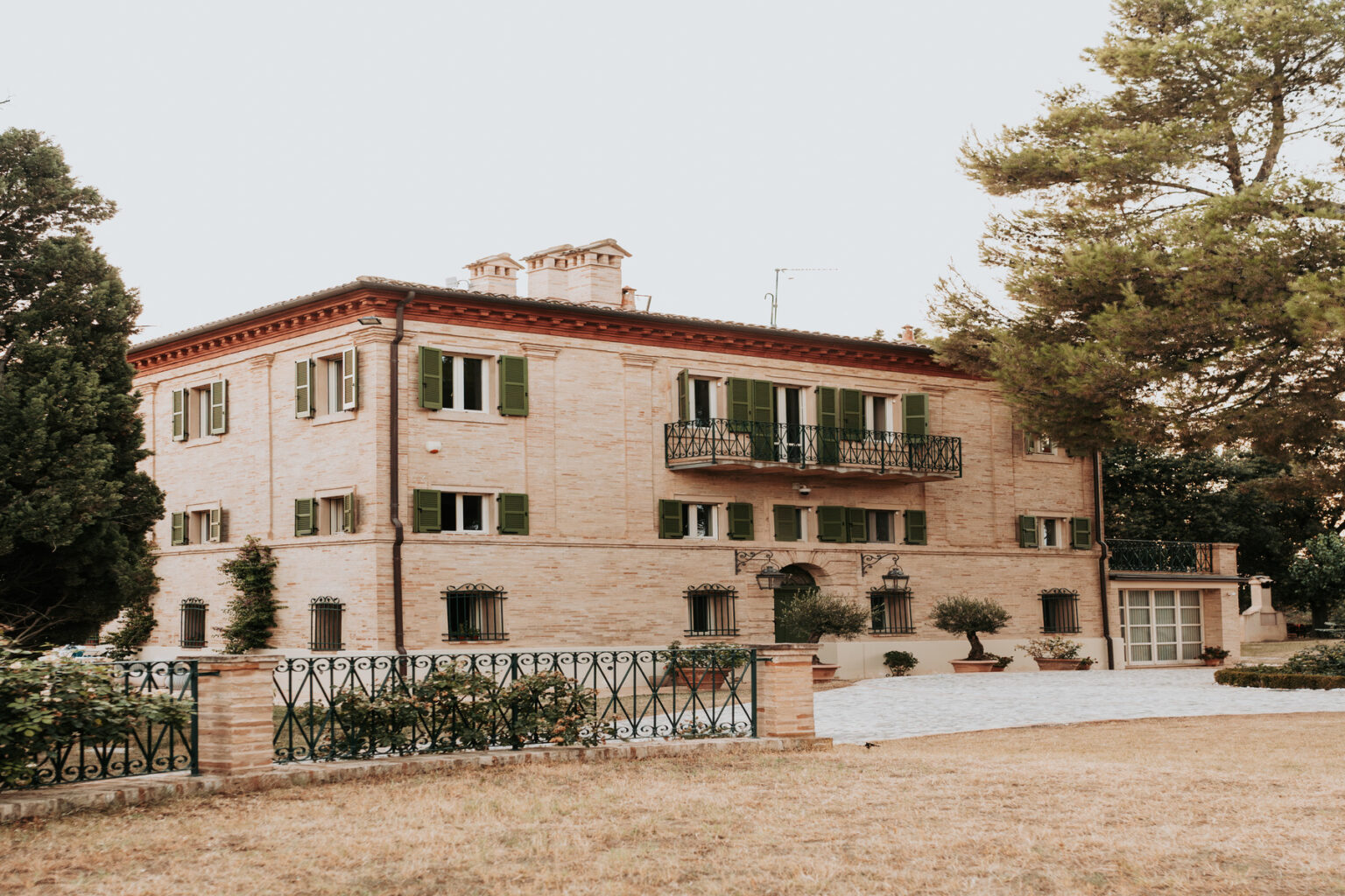 Elegant two-story brick house with green shutters, featuring a balcony and landscaped garden. The property includes a stone pathway and decorative plants, set against a serene backdrop. Ideal for showcasing architectural beauty and outdoor living spaces. | Only on SopranoVillas