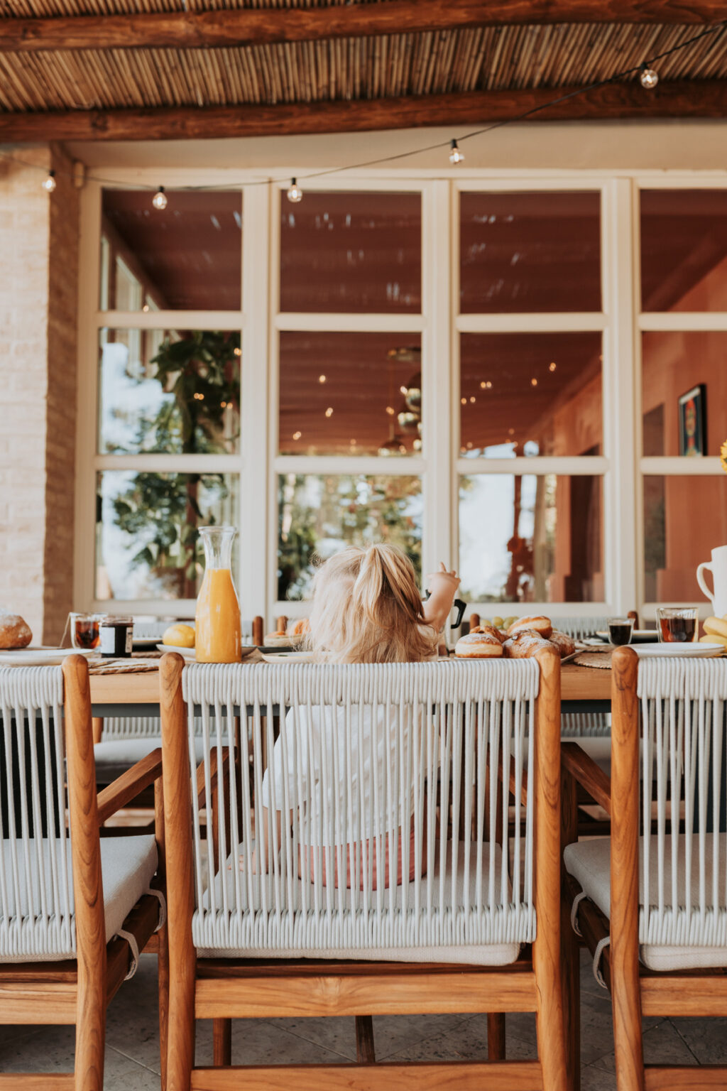 Child sitting at a breakfast table with a variety of food items, including pastries and orange juice, in a bright, airy dining space with large windows. | Only on SopranoVillas