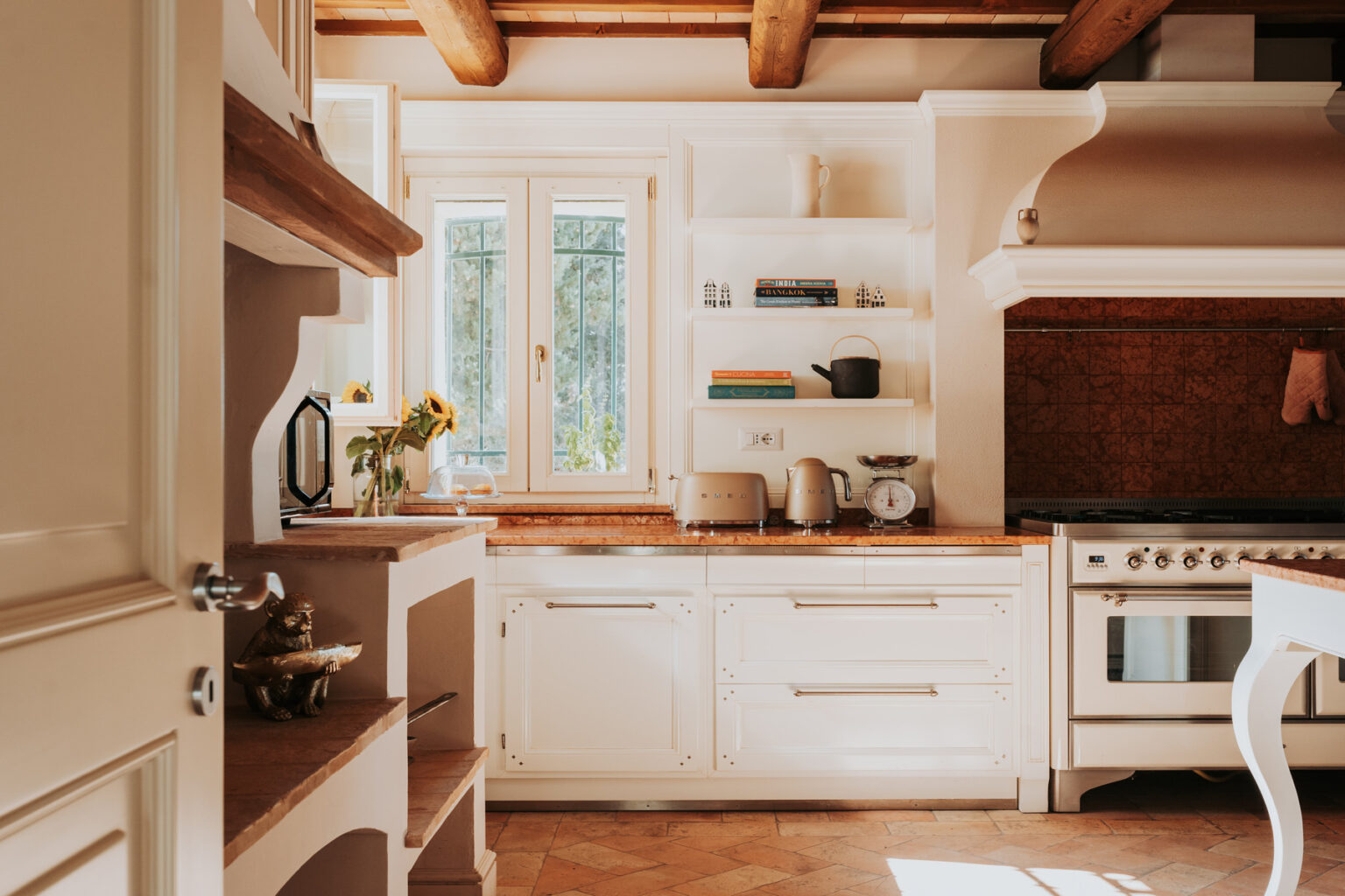 Bright and airy kitchen featuring white cabinetry, open shelves with kitchenware, and a rustic wooden countertop. Large window allows natural light to illuminate the space, enhancing its warm and inviting atmosphere. | Only on SopranoVillas