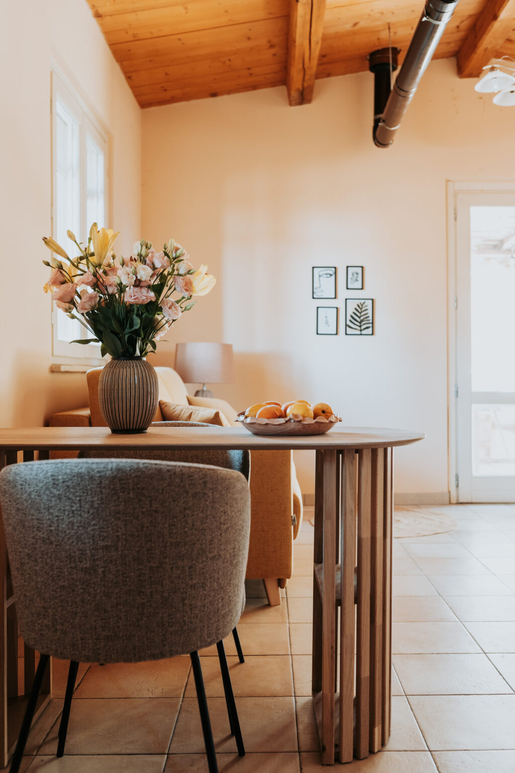 Cozy living room interior featuring a wooden dining table with a bowl of fresh fruits, a vase of colorful flowers, and framed wall art. Soft natural light illuminates the space, creating a warm and inviting atmosphere. | Only on SopranoVillas