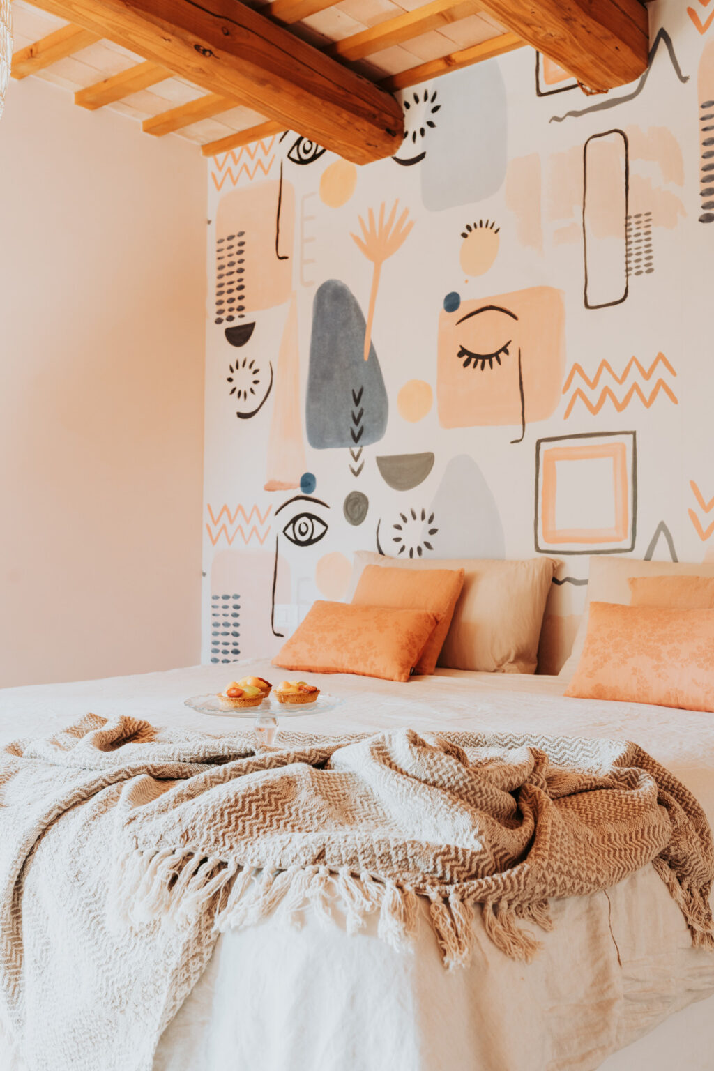 Cozy bedroom featuring a modern artistic mural with abstract shapes and patterns. The bed is adorned with soft beige and orange pillows, complemented by a textured throw blanket. A glass plate with pastries sits on the bed, enhancing the inviting atmosphere. | Only on SopranoVillas