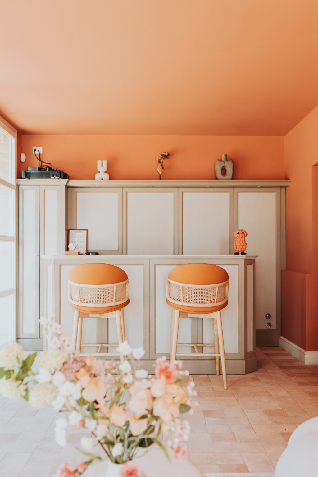 Modern home bar with orange walls and decor, featuring two stylish bar stools with orange cushions, a decorative shelf with unique ornaments, and a vase of fresh flowers in the foreground. | Only on SopranoVillas