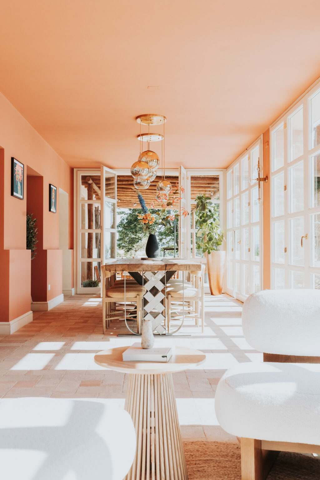 Bright and airy dining area with peach-colored walls, featuring a wooden table adorned with a vase of flowers. Large glass doors open to a sunny outdoor space, complemented by modern light fixtures and stylish furniture. | Only on SopranoVillas
