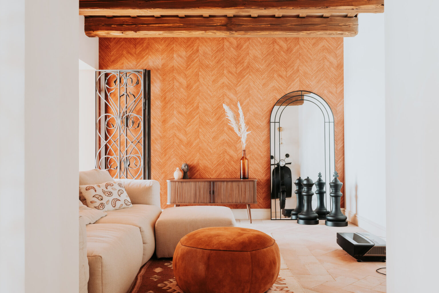 Modern living room featuring a beige sectional sofa with decorative pillows, a stylish orange herringbone accent wall, and a round suede ottoman. The room includes a wooden sideboard, a tall mirror framed in black, and decorative elements like a vase with pampas grass and oversized chess pieces, creating a cozy and inviting atmosphere. | Only on SopranoVillas