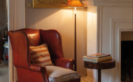 Cozy living room scene featuring a vintage red leather armchair with a striped cushion, a wooden side table stacked with books, and a warm floor lamp illuminating the space.