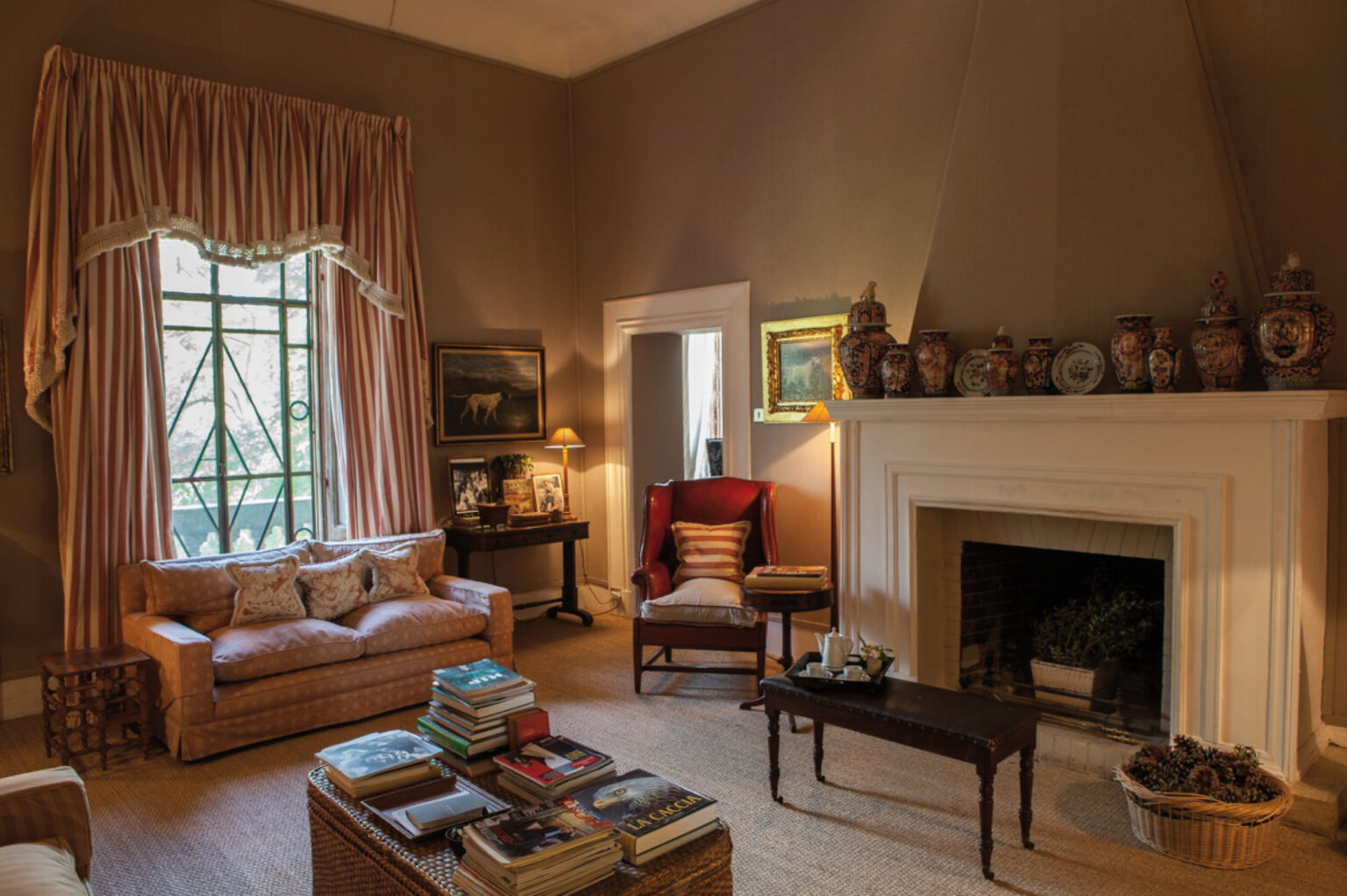 Cozy living room featuring a beige sofa, elegant curtains, a wooden coffee table stacked with books, and a fireplace adorned with decorative vases and artwork. Natural light streams through the window, enhancing the warm and inviting atmosphere. | Only on SopranoVillas