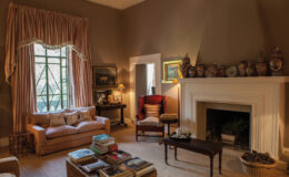 Cozy living room featuring a beige sofa, elegant curtains, a wooden coffee table stacked with books, and a fireplace adorned with decorative vases and artwork. Natural light streams through the window, enhancing the warm and inviting atmosphere.
