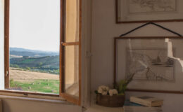 Cozy interior scene featuring an open window with a scenic view of rolling hills and fields. The room includes vintage framed maps on the wall, a white dresser with books and a decorative plant, and stylish lanterns on the floor, creating a warm and inviting atmosphere.