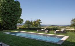 Modern outdoor swimming pool surrounded by lush greenery and lounge chairs, set against a scenic landscape with rolling hills and clear blue skies. Ideal for relaxation and leisure activities.