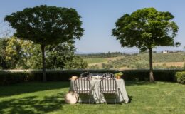 Outdoor dining setup in a scenic garden with two lush trees, overlooking a picturesque landscape of rolling hills and olive groves. Ideal for alfresco meals and gatherings.