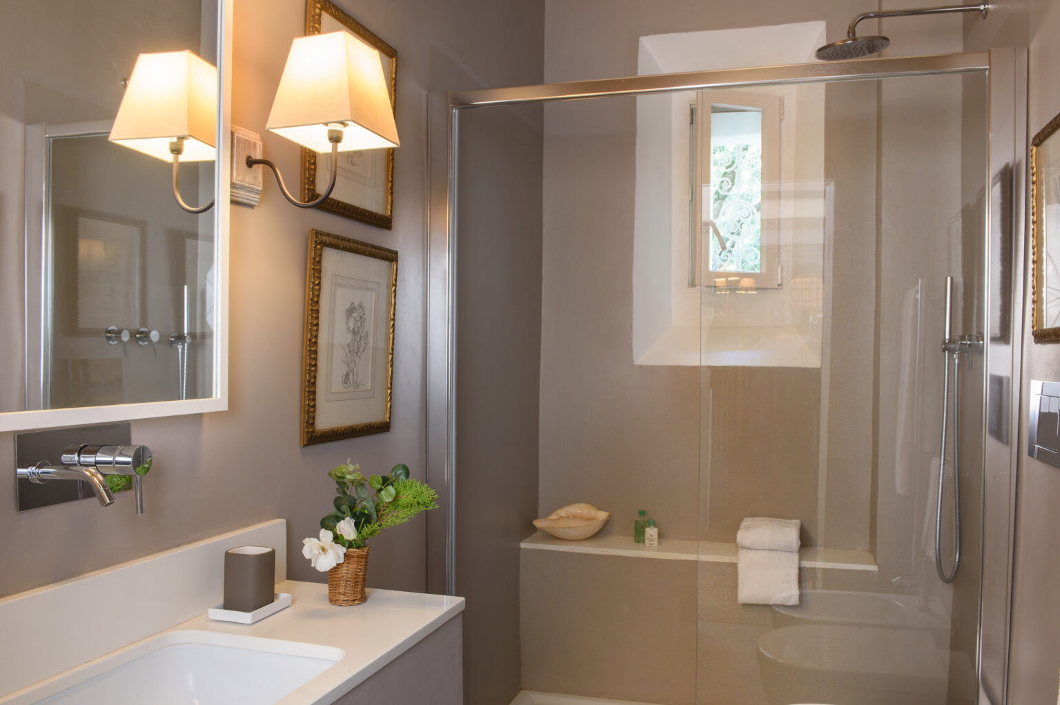 Modern bathroom interior featuring a glass shower, elegant wall sconces, and a stylish vanity with a small vase of flowers. The neutral color palette creates a serene atmosphere, complemented by framed artwork on the walls. | Only on SopranoVillas