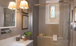 Modern bathroom interior featuring a glass shower, elegant wall sconces, and a stylish vanity with a small vase of flowers. The neutral color palette creates a serene atmosphere, complemented by framed artwork on the walls.