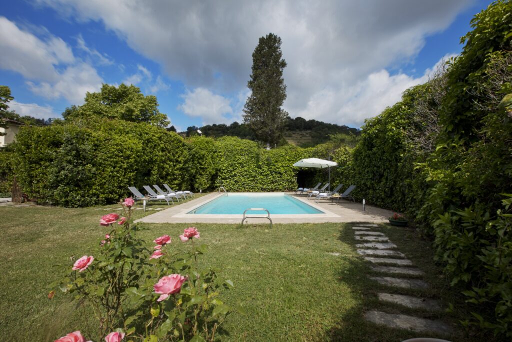 Serene backyard with a swimming pool surrounded by lush greenery and rose bushes, featuring lounge chairs and an umbrella for relaxation under a partly cloudy sky. | Only on SopranoVillas