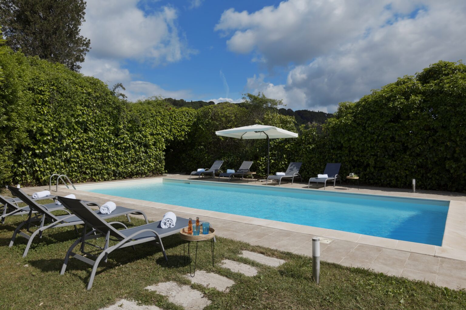 Outdoor swimming pool surrounded by lush greenery, featuring sun loungers, an umbrella, and a serene blue sky. Perfect for relaxation and leisure activities. | Only on SopranoVillas