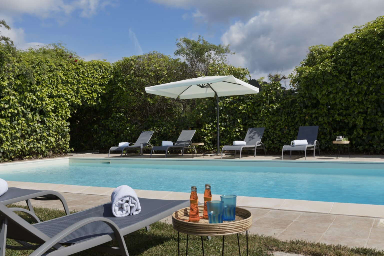 Luxurious outdoor pool area featuring comfortable lounge chairs, a shaded umbrella, and refreshing drinks on a table, surrounded by lush greenery and a clear blue sky. Perfect for relaxation and leisure. | Only on SopranoVillas