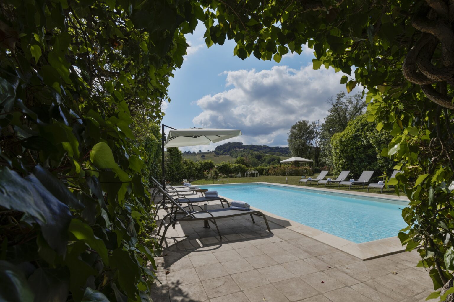 Serene swimming pool surrounded by lush greenery and lounge chairs, under a bright blue sky with fluffy clouds. Ideal for relaxation and leisure in a tranquil outdoor setting. | Only on SopranoVillas