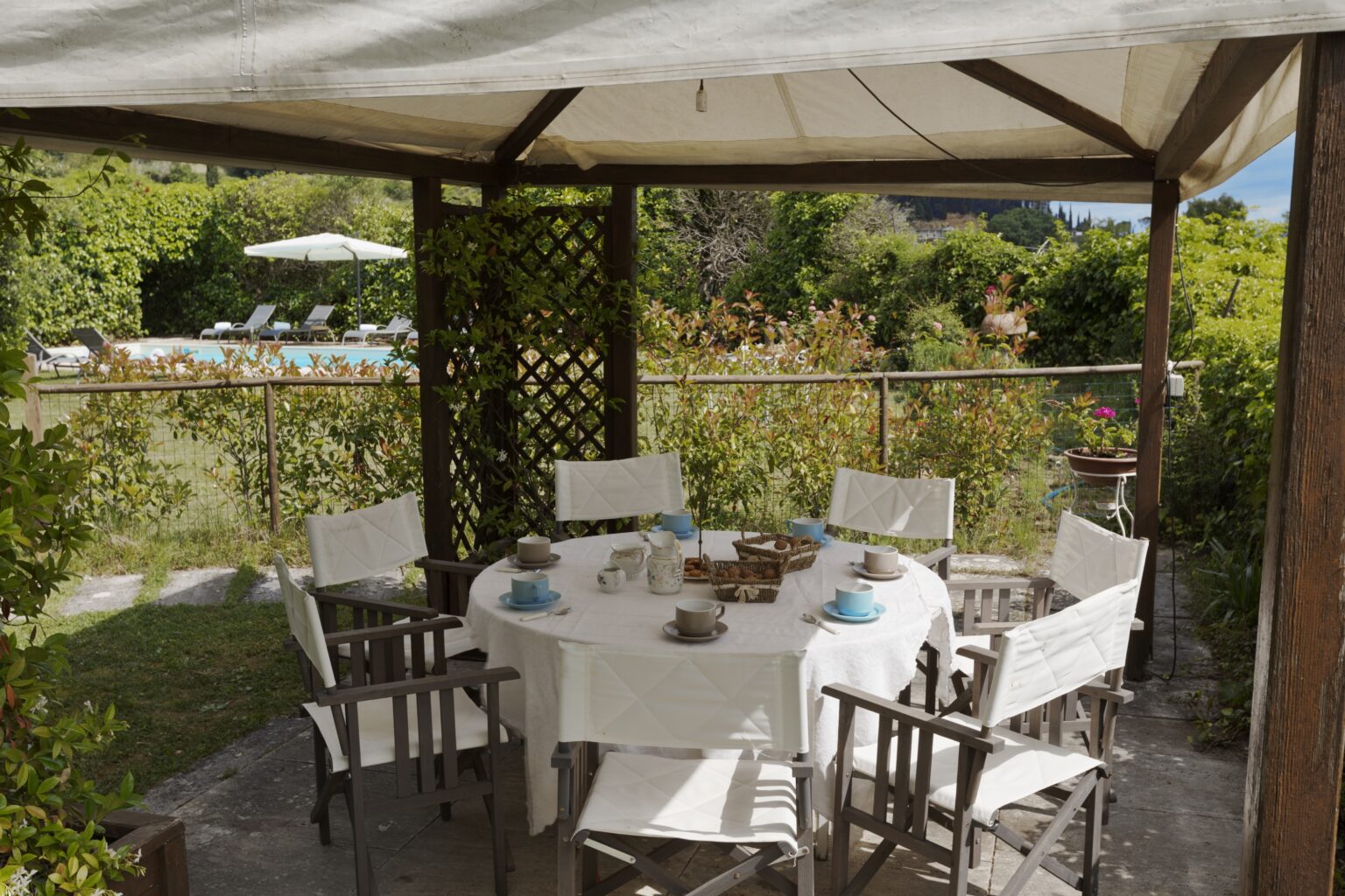 Outdoor dining area under a wooden pergola, featuring a round table set with blue dishes and surrounded by white chairs, with lush greenery and a swimming pool in the background. | Only on SopranoVillas