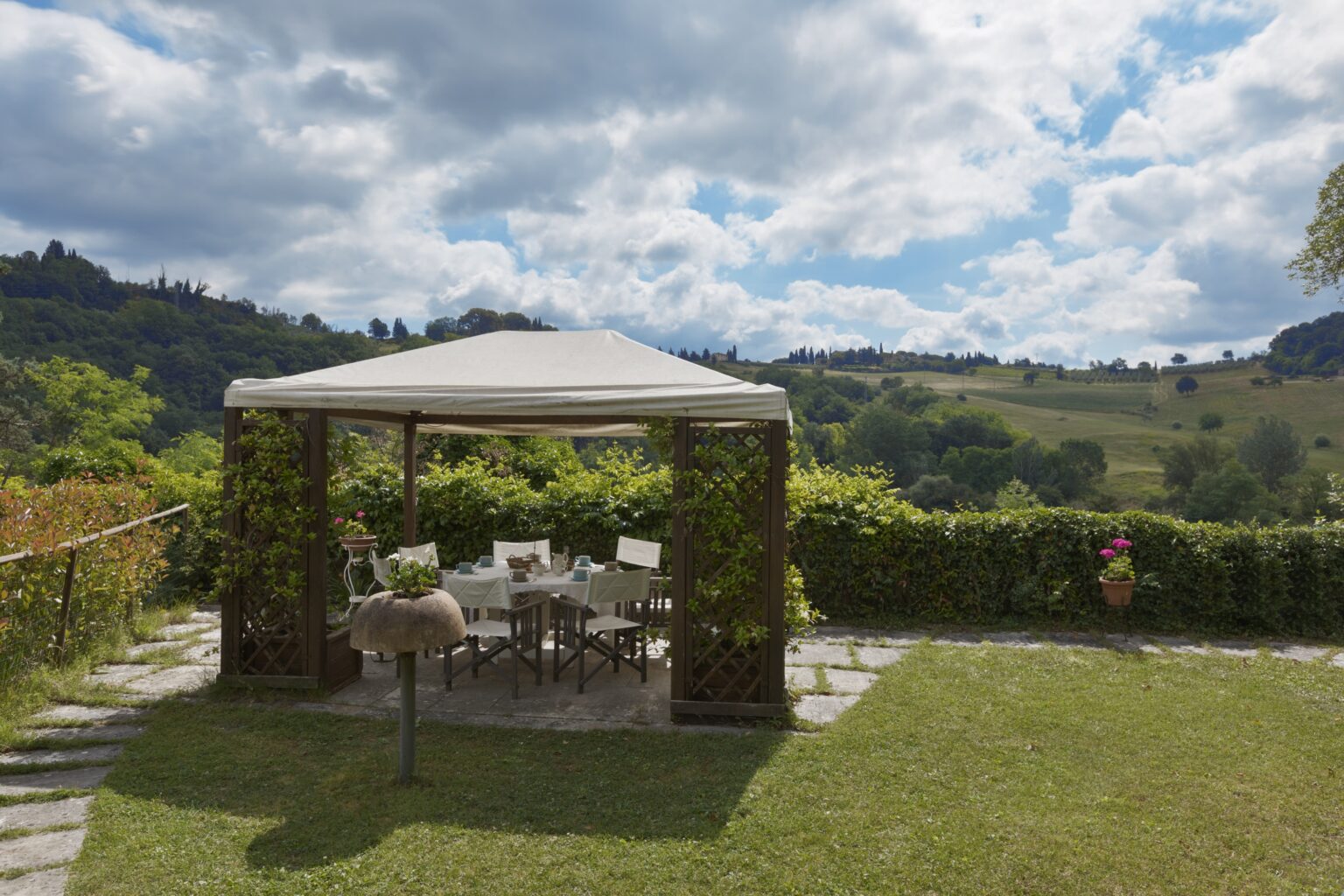 Outdoor dining area under a white gazebo surrounded by lush greenery and rolling hills, with a bright blue sky and fluffy clouds in the background. Ideal for relaxation and gatherings. | Only on SopranoVillas