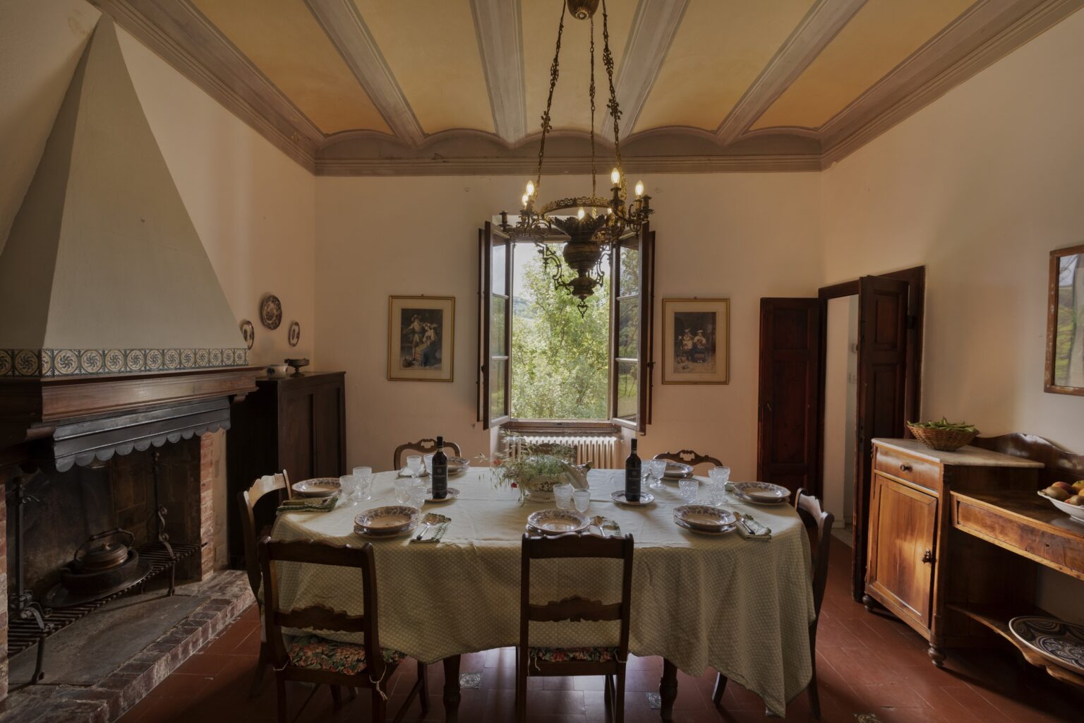Charming dining room featuring a large wooden table set for a meal, traditional decor including a vintage chandelier and a brick fireplace, with a view of greenery through open windows. | Only on SopranoVillas