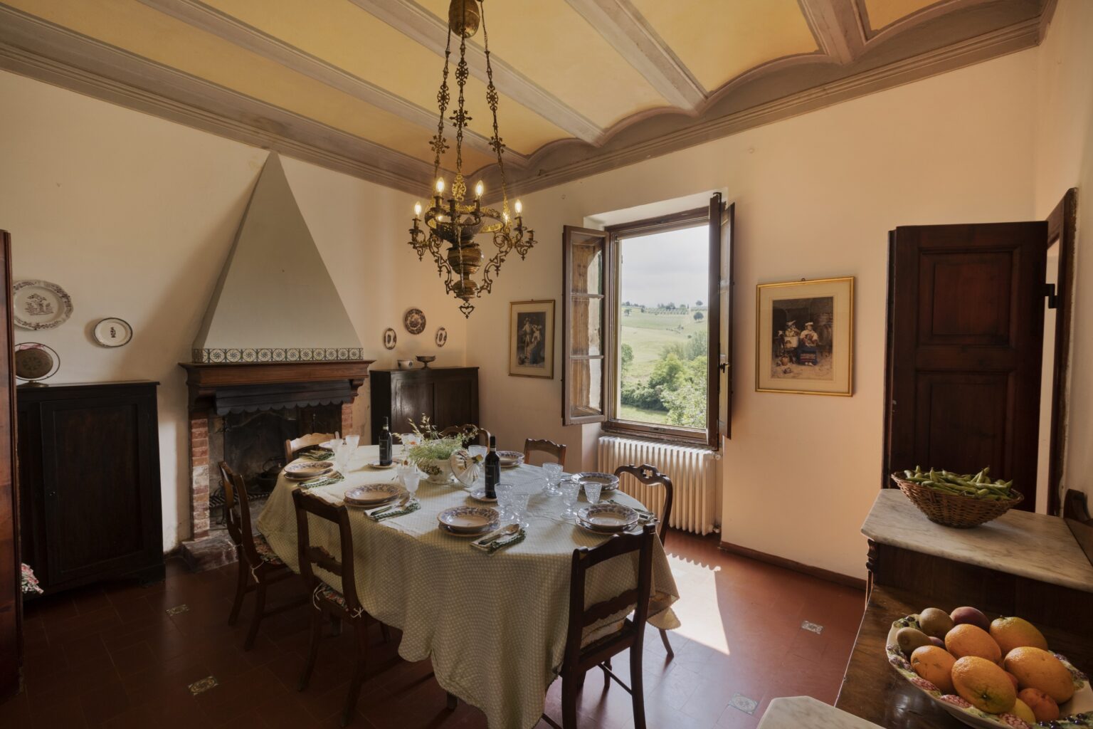 Elegant dining room with a rustic charm, featuring a large table set for a meal, a chandelier, and a scenic view through open windows. The room includes a fireplace, wooden furniture, and decorative wall art, creating a warm and inviting atmosphere. | Only on SopranoVillas