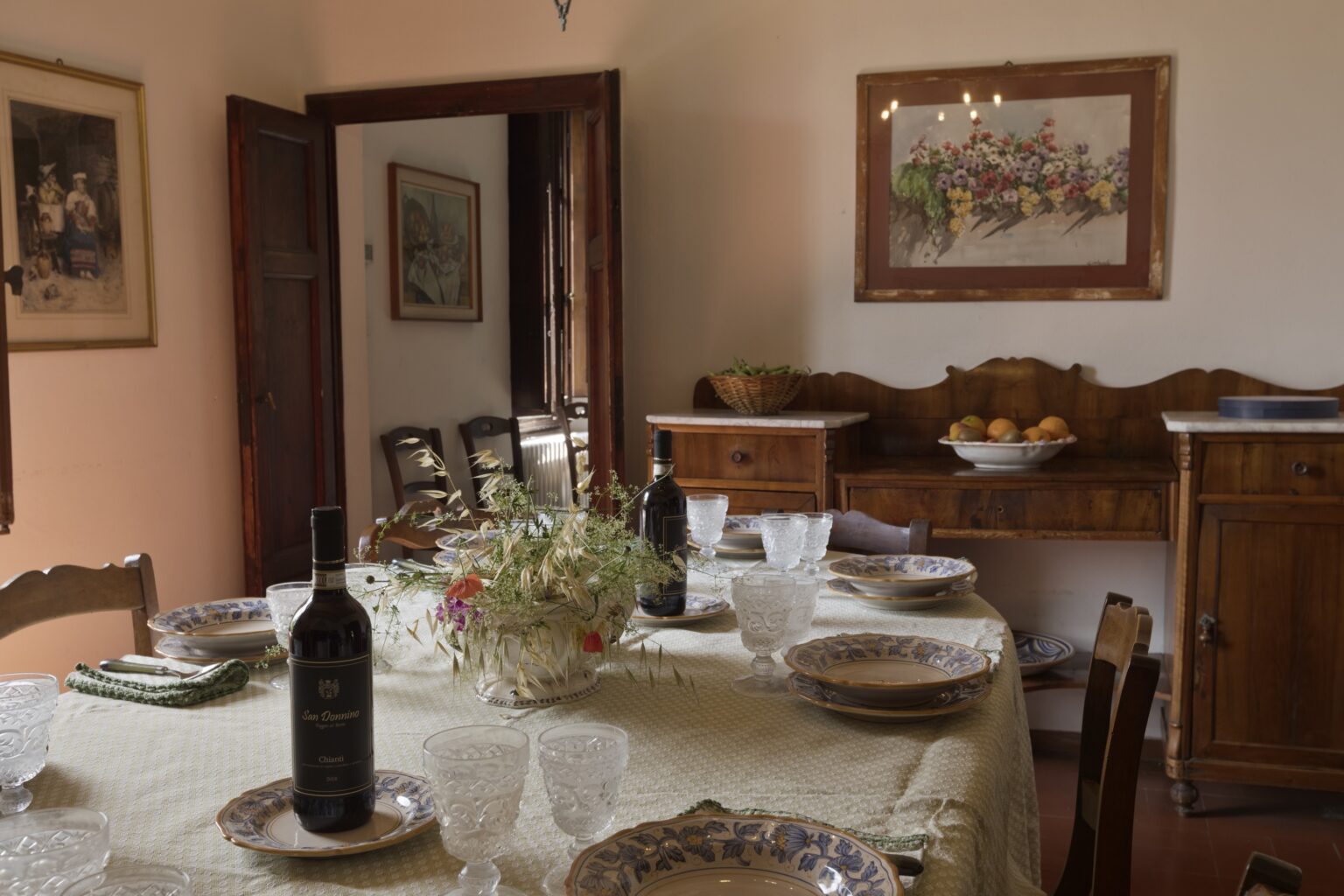 Cozy dining room setup featuring a beautifully arranged table with plates, glasses, and a bottle of wine, complemented by floral decor and wooden furniture, creating an inviting atmosphere for gatherings. | Only on SopranoVillas