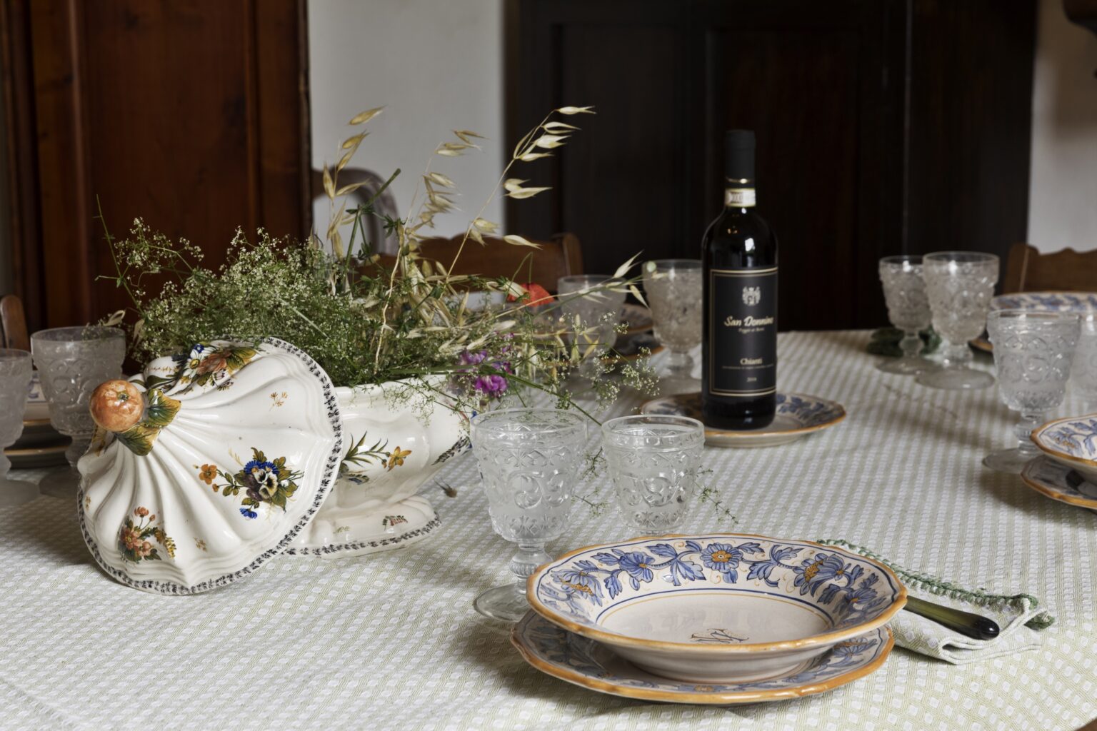 Elegant dining table set with vintage china plates, crystal glassware, and a decorative floral centerpiece, accompanied by a bottle of red wine. Perfect for a festive gathering or special occasion. | Only on SopranoVillas