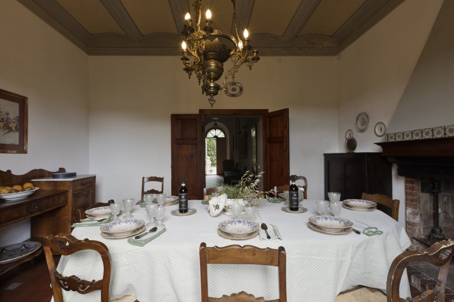 Elegant dining room featuring a beautifully set table with fine china, glassware, and decorative centerpieces, illuminated by a vintage chandelier. Wooden furniture and a cozy fireplace enhance the warm atmosphere of the space. | Only on SopranoVillas