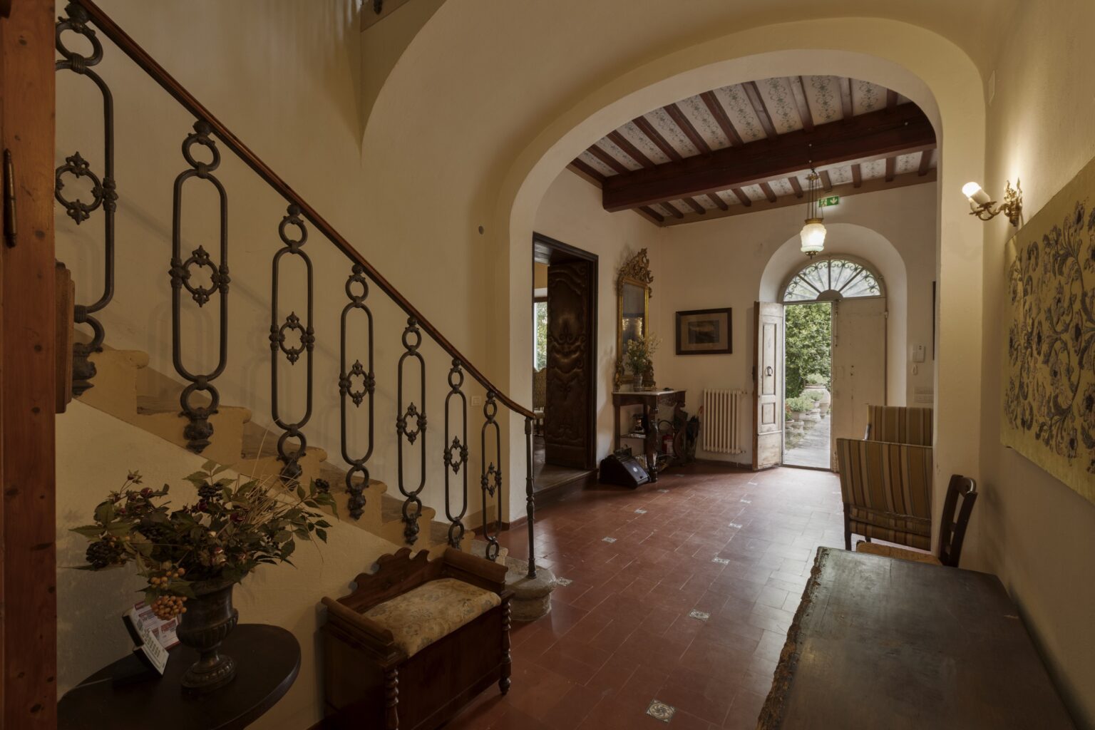 Elegant entryway featuring a wrought iron staircase, wooden accents, and a vintage decor. The space includes a decorative vase with foliage, a wooden bench, and an inviting open door leading to an outdoor garden. Natural light enhances the warm tones of the interior. | Only on SopranoVillas