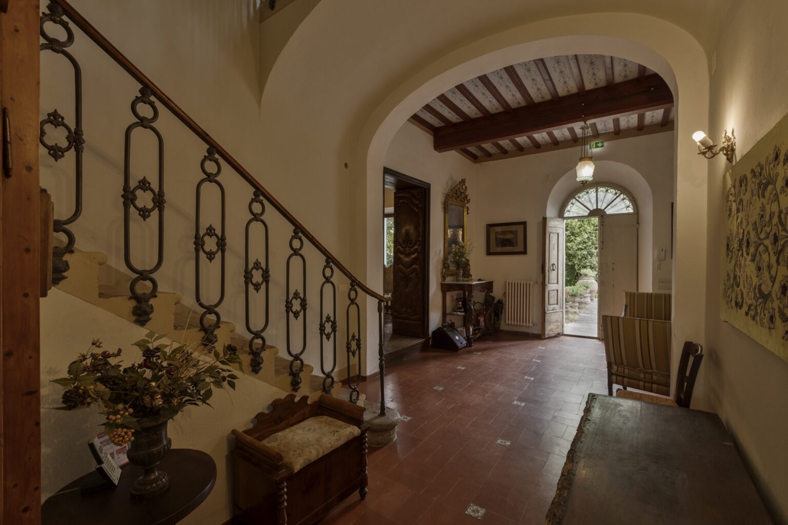 Interior view of a charming entrance hall featuring a decorative staircase with wrought iron railings, warm lighting, and rustic decor elements, leading to a door that opens to a garden area. | Only on SopranoVillas