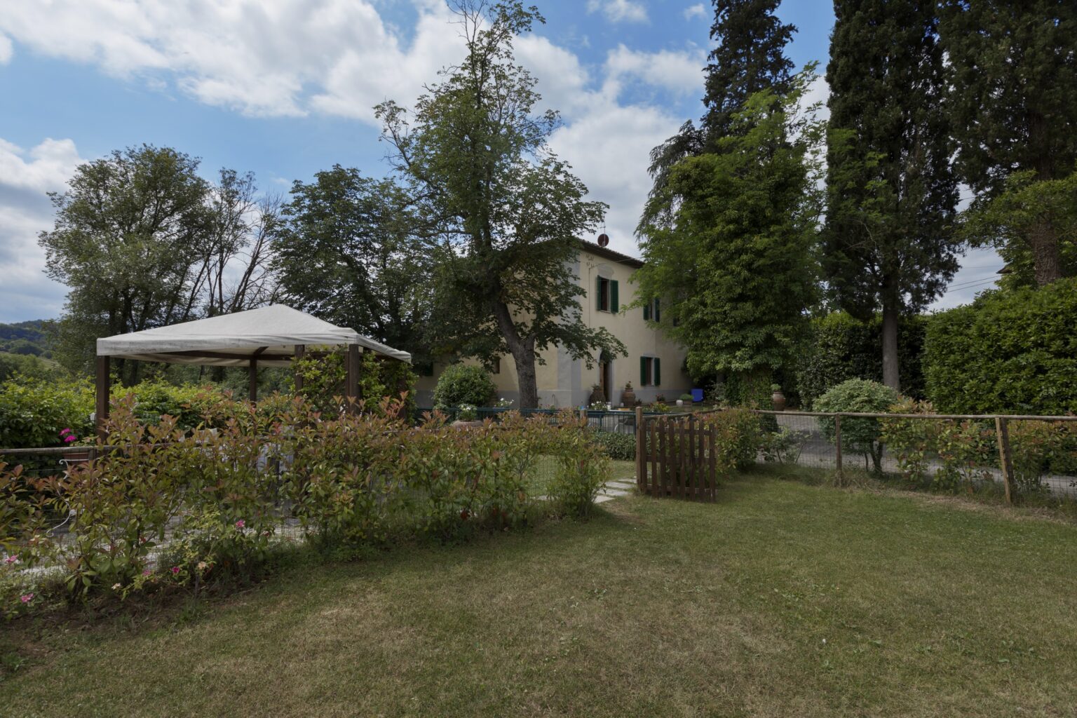 Charming countryside villa surrounded by lush greenery and a well-maintained garden, featuring a shaded patio area and beautiful landscaping under a partly cloudy sky. | Only on SopranoVillas