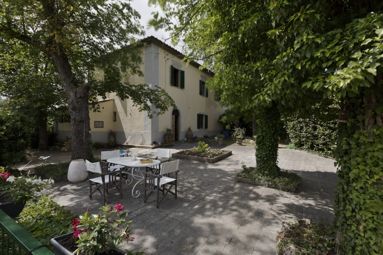 Charming outdoor patio area with a dining table and chairs surrounded by lush greenery and flowering plants, featuring a rustic house in the background. Ideal for relaxing and enjoying nature. | Only on SopranoVillas