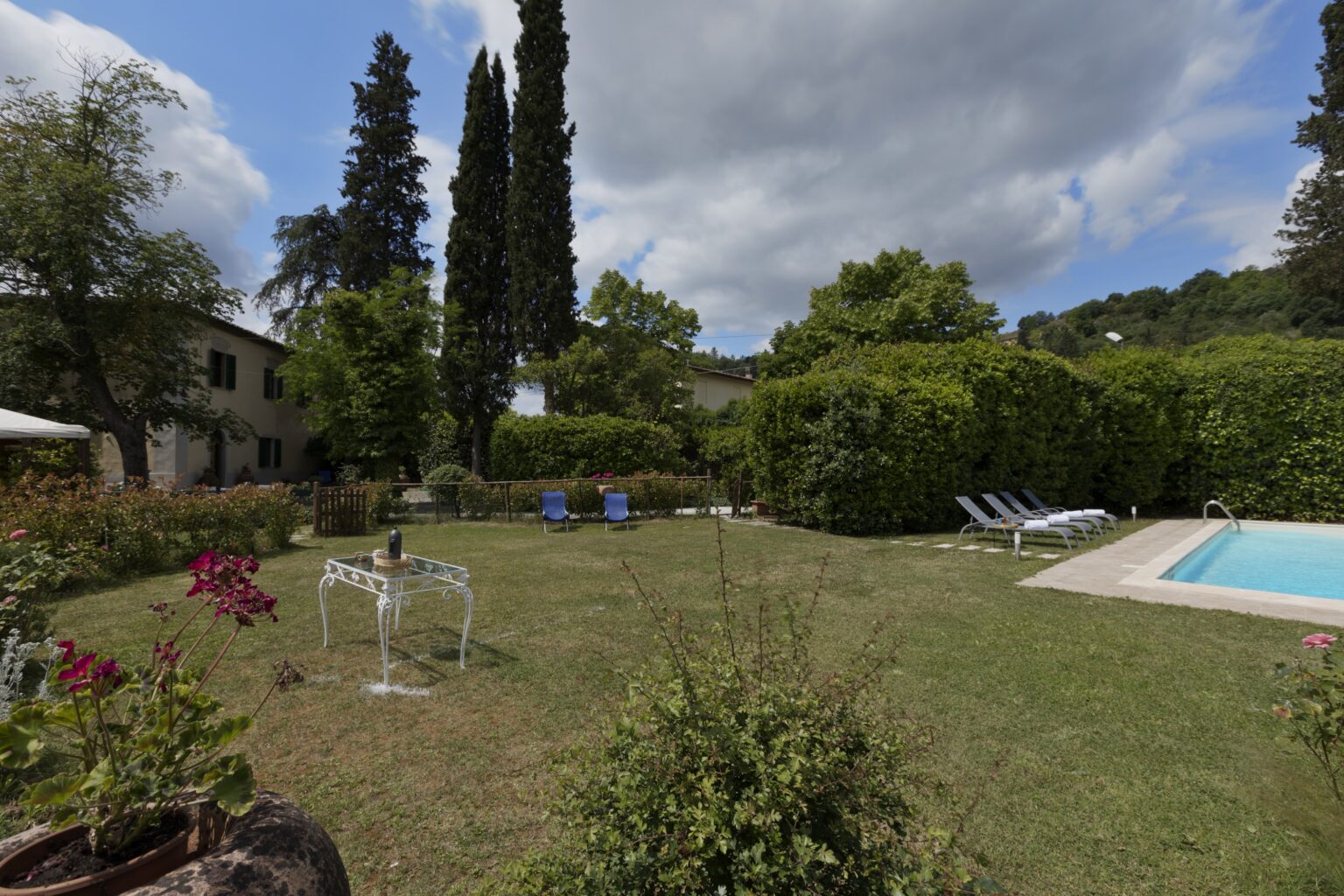 Scenic outdoor garden with lush greenery, vibrant flowers, and a swimming pool surrounded by lounge chairs, set against a backdrop of blue skies and clouds. | Only on SopranoVillas