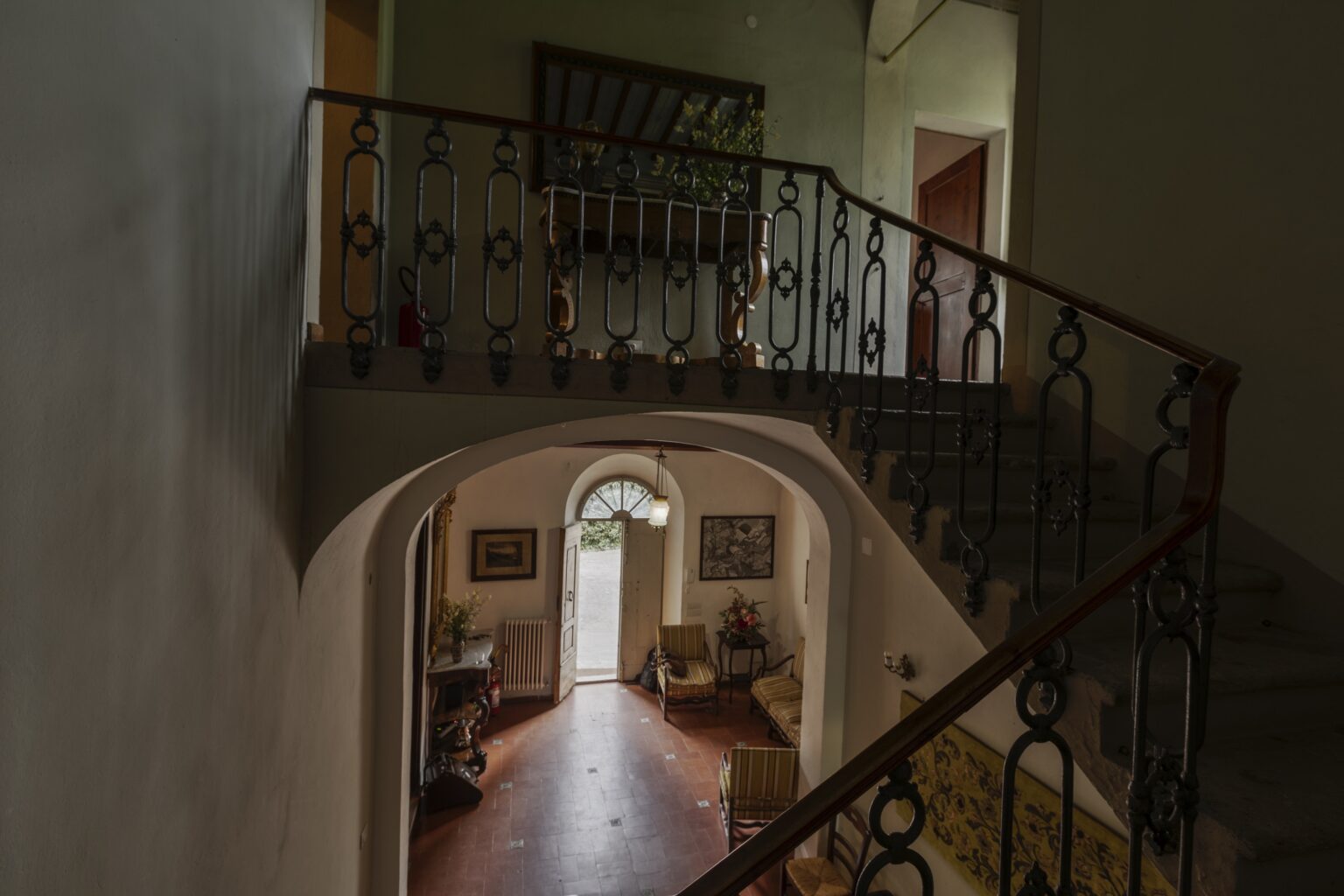 Interior view of a home showcasing a staircase with decorative wrought iron railing, leading to a well-lit hallway with an arched entrance, surrounded by cozy furnishings and artwork. | Only on SopranoVillas