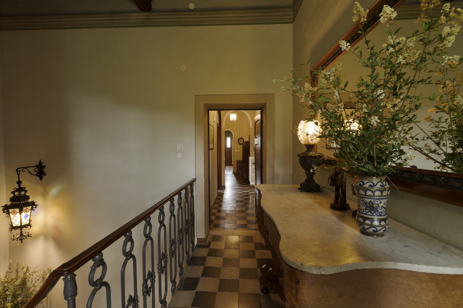 Elegant hallway featuring a vintage wooden console table with a marble top, adorned with decorative vases and lamps. The space is illuminated by a charming wall lantern, showcasing patterned floor tiles and a view into a well-lit corridor. | Only on SopranoVillas
