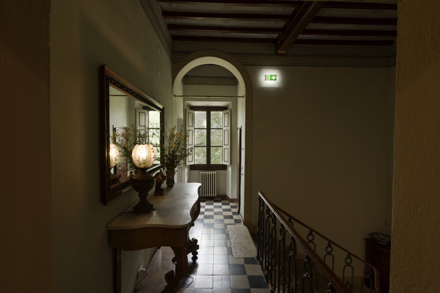 Elegant hallway with a wooden console table, decorative mirror, and natural light streaming through a large window, showcasing a classic interior design with checkered flooring and a staircase. | Only on SopranoVillas