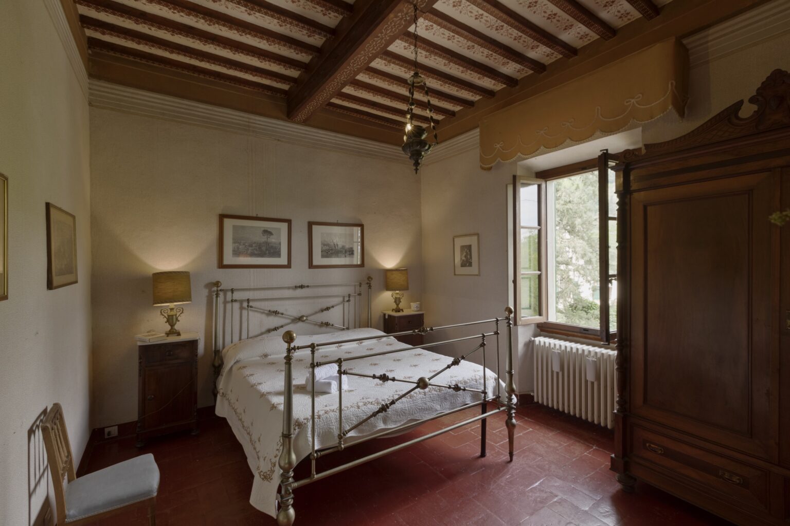 Cozy bedroom featuring a wrought iron bed, warm lighting from bedside lamps, and rustic wooden beams. The room is decorated with framed artwork, a vintage wardrobe, and a large window offering natural light and a view of greenery outside. | Only on SopranoVillas